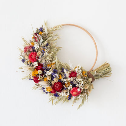 wood wreath hoop half covered in dried flowers. flowers red, white blue and yellow with pale green grasses and oats. The dried flower wreath is hung on a white wall