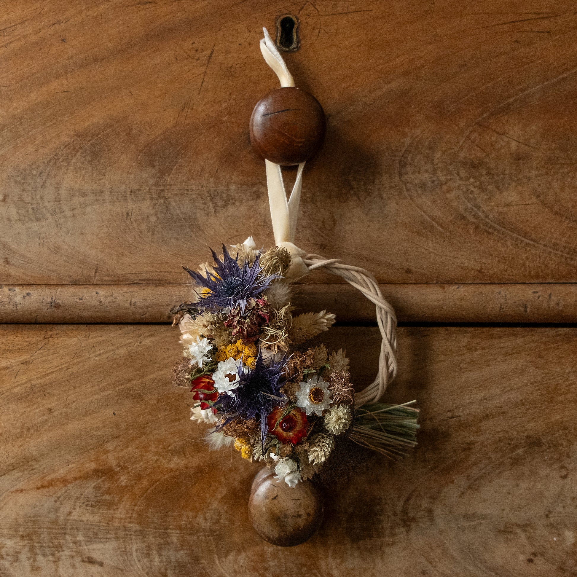 Decorative arrangement dried flower wreath half covered in flowers blue thistles, red zinnia and strawflowers, yellow lona and white winged daisies. the mini gift sized wreath is hung via a cream velvet ribbon on a old chest of drawer handle