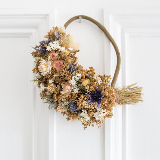 small willow wreath half covered in dried flowers. blue thistles, white strawflowers, golden broom bloom. the wreath is hung on the back of a white door