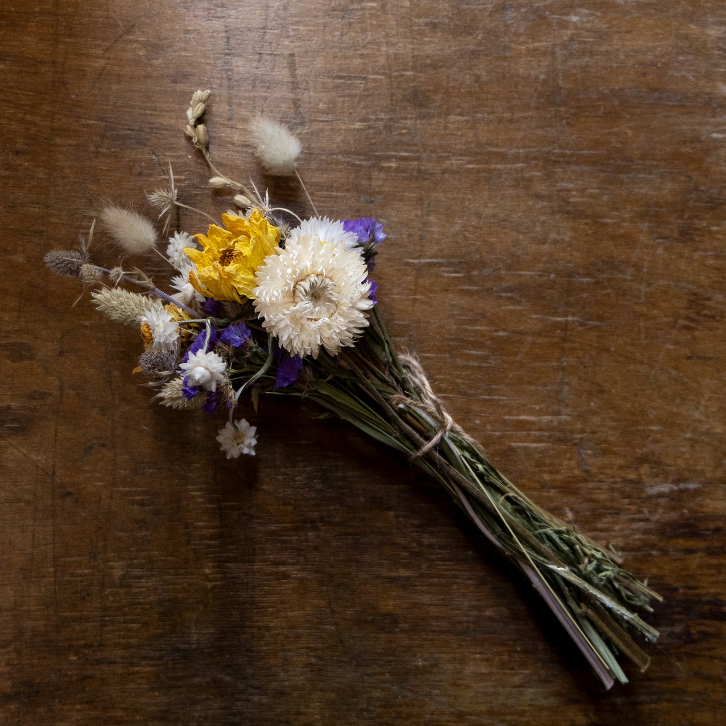 posy made of dried flowers on a wooden surface. white strawflowers, yellow dahlias and blue statice