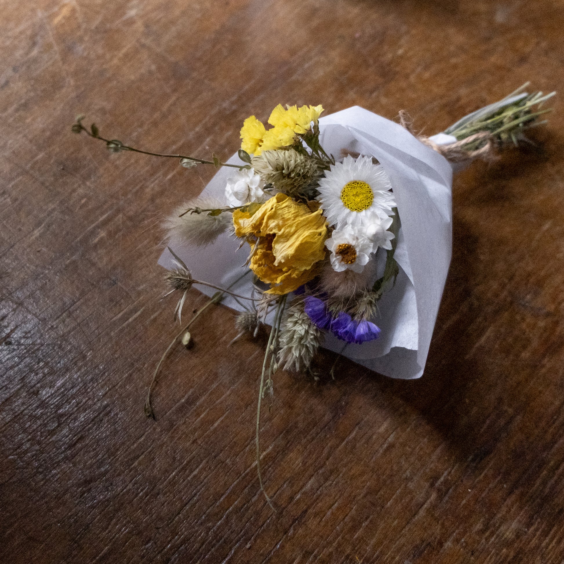 flower posy laid on a wooden table. the posy is made of yellow white and blue dried flowers with grasses
